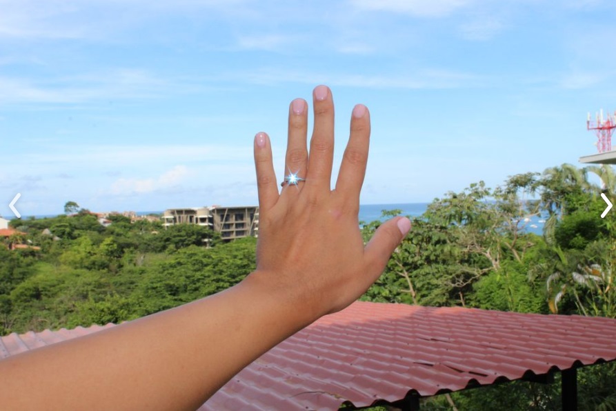 In this picture a girl is showing off her diamond engagment ring with the ocean in the background